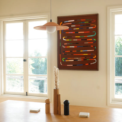 Wall art for dining room featuring a framed abstract wall art piece with multicolored curved lines on a dark brown background, hanging on a cream wall in a bright room with large windows, a wooden table, and a pendant light.