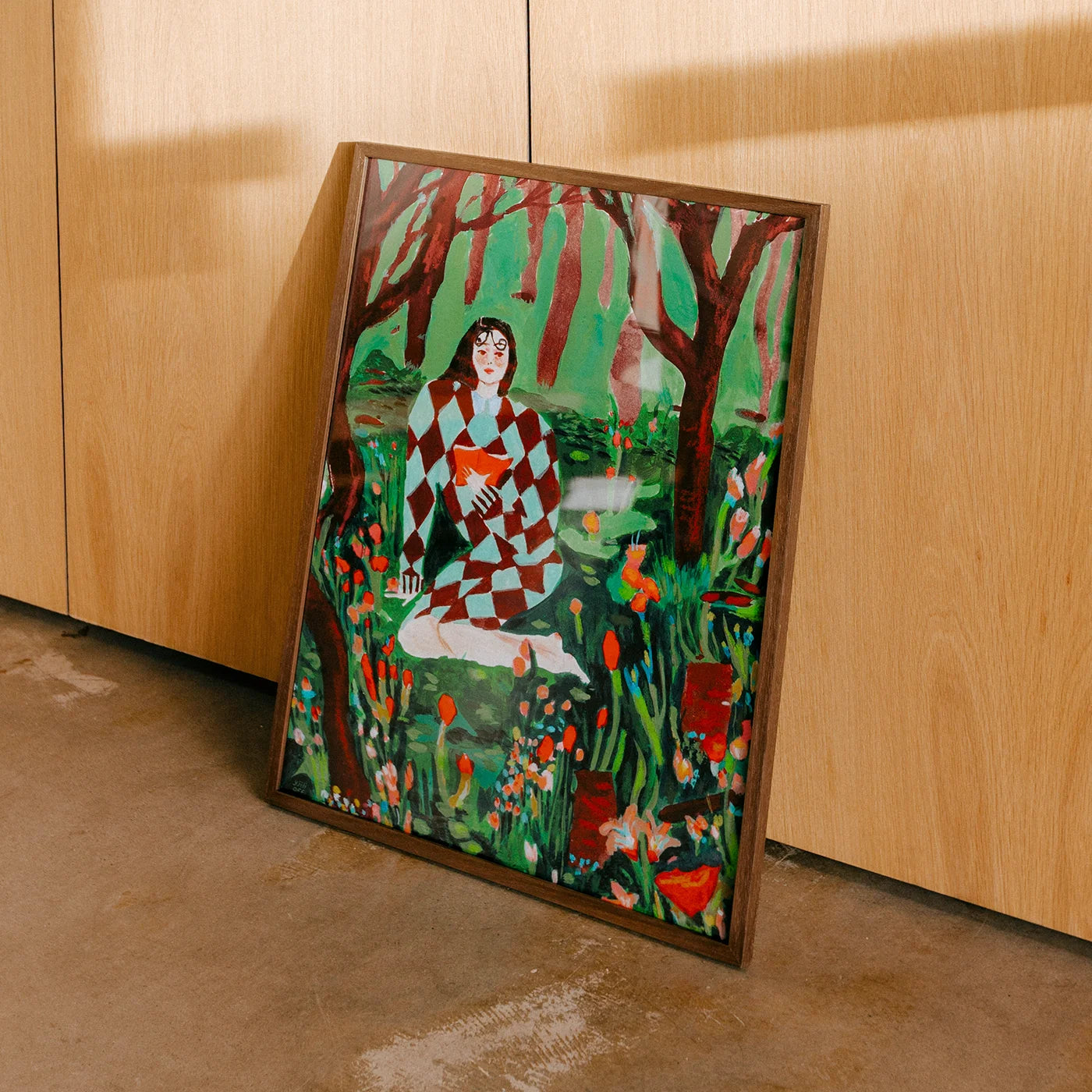 Framed wall art leaning against a light wood cabinet on a concrete floor. The art print depicts a seated woman in a red-and-white checkered dress surrounded by green foliage, red trees, and orange flowers.