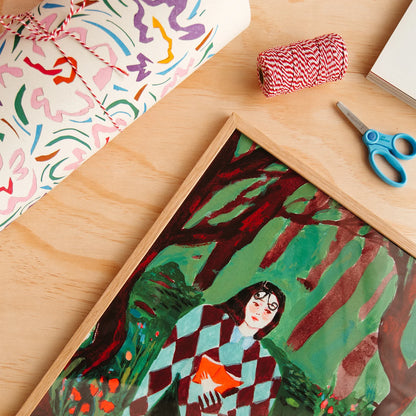 A framed art print depicting a woman in a red and white checkered coat holding an orange book in a forest, styled on a light wood table with a wrapped gift, bakers twine, and blue-handled scissors.