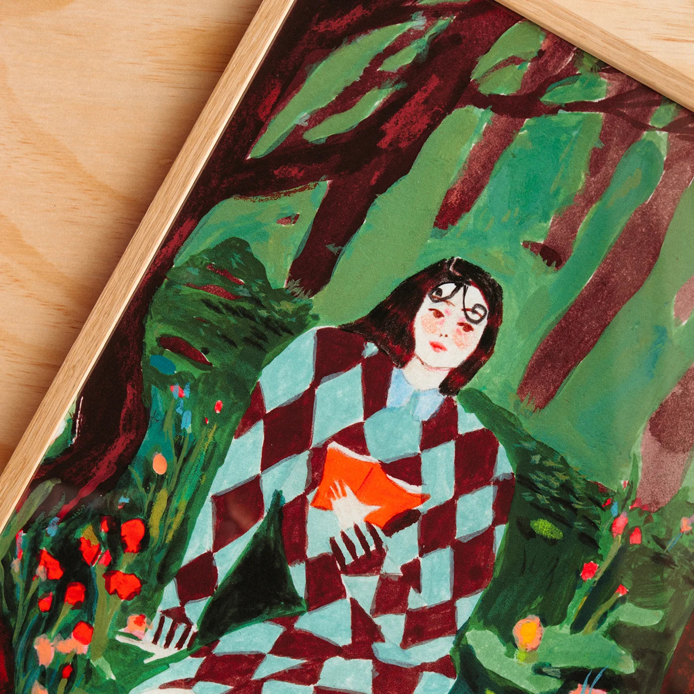 A framed art print of a woman in a checkered red and light blue dress sitting in a forest, holding an orange book, displayed on a light wood surface. The wall art has a watercolour look.