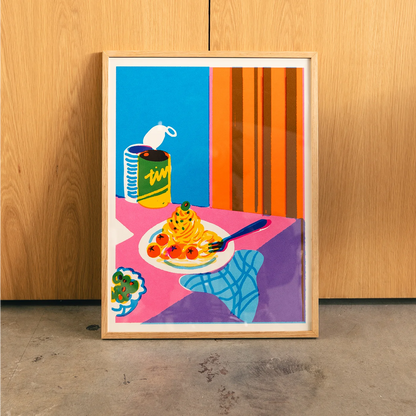 A framed pop art print depicting a colorful still life of spaghetti, tomatoes, broccoli, and a can of soup, leaning against a light wood-paneled wall on a concrete floor.