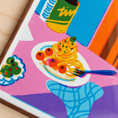 A close-up of a colorful pop-art style print showing a plate of spaghetti with tomatoes, a fork, a can, and bold geometric shapes in pink, blue, purple, orange, and yellow. Framed in walnut timber.
