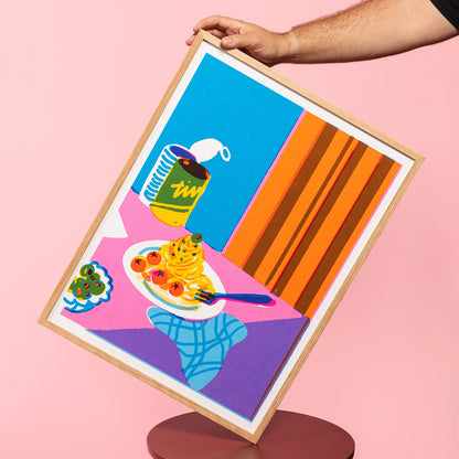 A hand holds a light wood framed art print against a solid pink background. The print features a colorful pop-art illustration of a breakfast table scene with a plate of spaghetti and tomatoes, a blue can, and bold geometric shapes in blue, pink, orange, and purple.