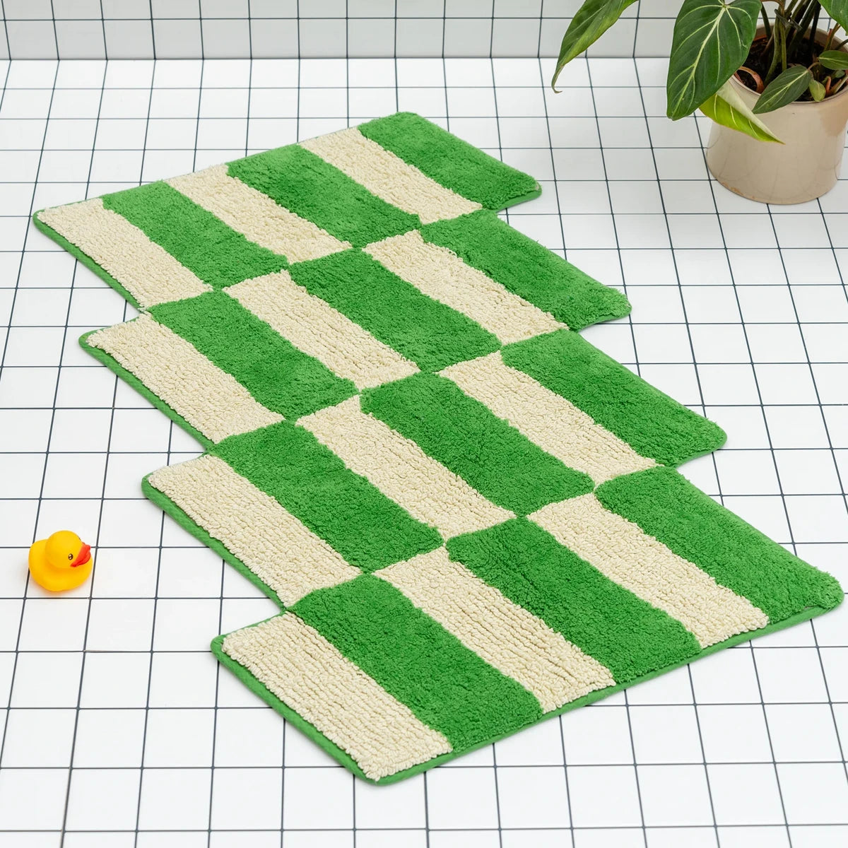 Green and beige checkered bath mat by Tarta Gelatina on a grid background with a plant and rubber duck.