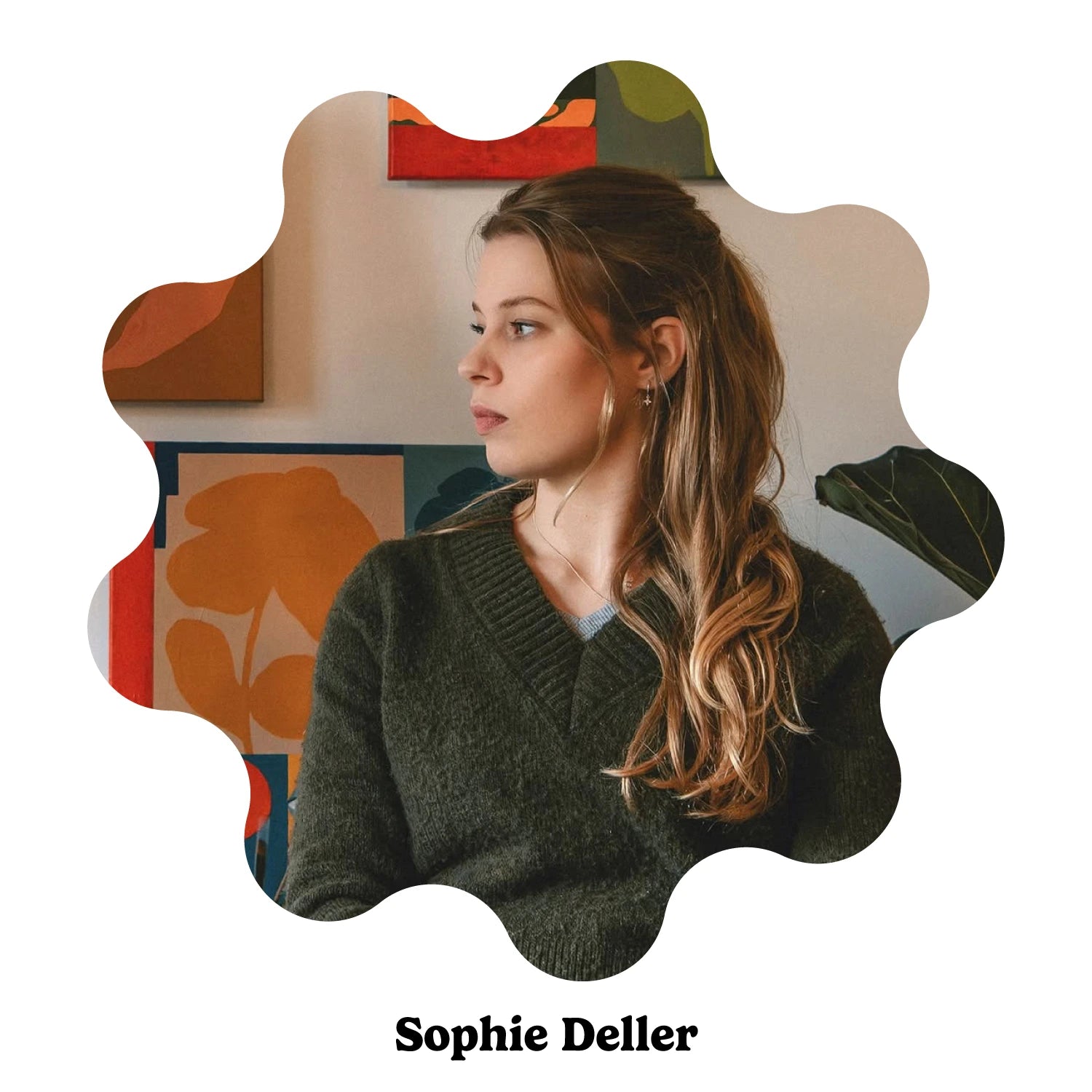 Portrait of English artist Sophie Deller sitting in a wooden chair wearing a dark olive green knit sweater, set against a cozy room decorated with colorful floral paintings and a potted plant.